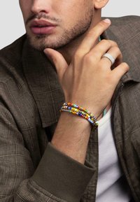 Colorful bead bracelet with multiple strands in red, yellow, blue, and green. Worn on a hand with a silver ring, dressed in a patterned brown jacket.