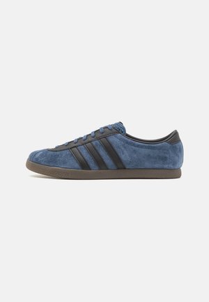 Niskie sneakersy z niebieską, zamszową cholewką, czarnymi skórzanymi paskami i zakładką na pięcie, niebieskimi sznurowadłami oraz brązową gumową podeszwą, widoczne z profilu bocznego.