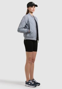 Jeune femme debout portant une veste matelassée grise, une casquette noire, un short noir et des baskets bleu marine sur un fond gris uni.