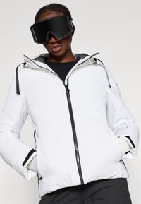 Fehér puffermellény cipzáras elejével, állítható kapucnival és fekete részletekkel. Textúrált felülettel és elasztikus mandzsettákkal rendelkezik.