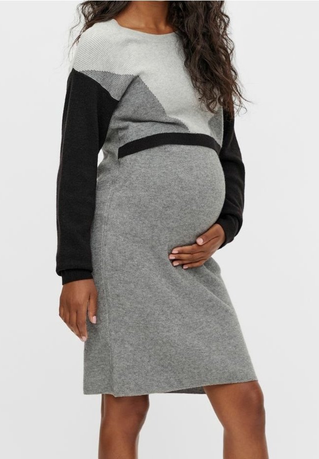 Femme enceinte portant une robe en maille à manches longues grise et noire, tenant doucement son ventre d'une main.