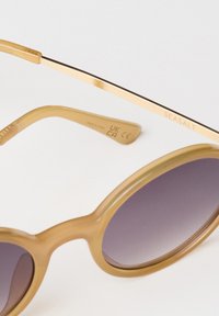 Ochelari de soare ovulari cu nuanțe de maro și lentile întunecate. Rame transparente cu o textură netedă, având un accent din metal de culoare aurie pe tâmple.