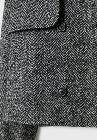 Manteau à motif en chevrons noir et blanc avec deux boutons noirs et un rabat de poche visible sur un fond neutre.