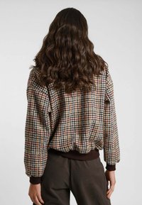 Donna con capelli castani ondulati che indossa una giacca a fantasia check con polsini e vita a costine, girata di spalle su uno sfondo neutro.