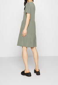 Olive zelené šaty s krátkým rukávem a vrstvenou sukní. Vyrobené z jemného materiálu, mají kulatý výstřih. Nosič má na sobě černé platformové sandály.
