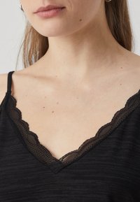 Černý bezrukávový top s výstřihem do V a krajkovým lemem s vlnkami, texturovaná tkanina a tenká ramínka.