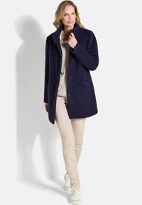 Marineblauer Wollmischungsmantel mit Kragen, vorderen Knöpfen und zwei Seitentaschen. Kombiniert mit beigen Hosen und weißen Turnschuhen.