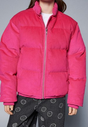 Person iført en klar pink fløjlsvelour-fyldt jakke med lynlås foran, kombineret med sorte bukser med smiley-mønstre.
