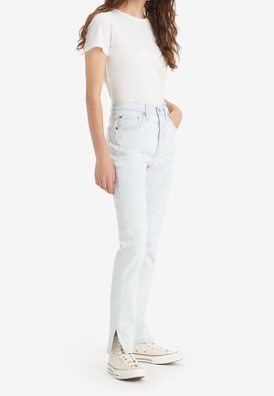 Vrouw draagt een wit T-shirt met korte mouwen, lichtblauwe high-waist jeans met een voorste split aan de zoom en witte hoge sneakers.