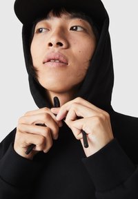 Jeune personne portant un sweat à capuche noir et une casquette, ajustant les cordons de la capuche en regardant légèrement vers le haut et sur le côté.