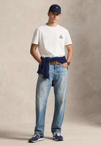 Λευκό βαμβακερό t-shirt με λογότυπο, μπλε τζιν, ναυτικό πουλόβερ δεμένο γύρω από τη μέση, ναυτικό καπέλο, μπλε αθλητικά παπούτσια με λευκές λεπτομέρειες, ρολόι χειρός.