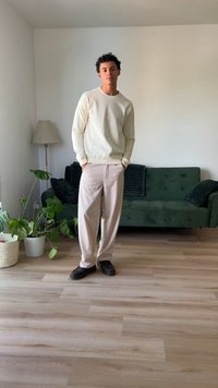 Maglione chiaro, pantaloni beige a gamba larga e scarpe scure. In piedi in una stanza con un divano verde, una lampada e una pianta in vaso su un pavimento di legno.
