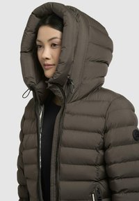 Brown puffer jacket with a large hood, zip closure, and ribbed texture. Features adjustable drawstrings and a black logo patch on the sleeve.