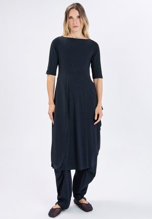 Donna con capelli castano chiaro che indossa un lungo vestito nero con maniche fino al gomito e scarpe nere basse, in piedi davanti a uno sfondo bianco.