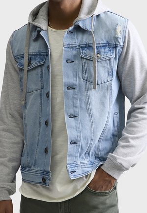 Man draagt een lichtblauwe spijkerjack met grijze sweatshirtsleeves en capuchon, een beige overhemd eronder, en heeft zijn hand in de zak van een groene broek.