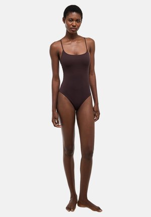 Femme grande aux cheveux courts, debout pieds nus, portant un maillot de bain une pièce brun foncé avec de fines bretelles, sur un fond uni.