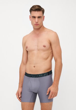 Graue Boxer-Briefs aus dehnbarem Stoff, mit einem schwarzen Taillenbund, auf dem "UNDER ARMOUR" in blauer Schrift steht. Eng anliegende Form, glatte Textur.