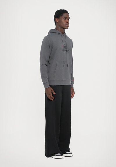 Grauer Kapuzenpullover mit Fronttasche, Kordelzug und Logodetail. Kombiniert mit lockeren schwarzen Hosen und schwarz-weißen Turnschuhen.