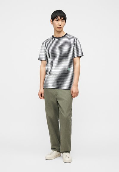 Νεαρός άνδρας που στέκεται, φοράει κοντομάνικο πουκάμισο με ασπρόμαυρες ρίγες, πράσινα παντελόνια και λευκά αθλητικά παπούτσια σε απλό φωτεινό φόντο.