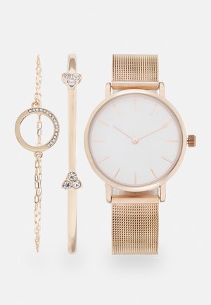 Gouden mesh horloge met witte wijzerplaat en roségouden accenten, vergezeld van twee gouden armbanden, waarvan één met heldere kristallen en één met een cirkelvormig ontwerp.