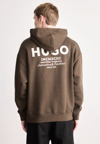 Ruda džemperis su gobtuvu, kurio priekyje yra didelis baltas užrašas „HUGO“ ir prekės ženklo detalės gale. Minkšta tekstūra ir laisvas siluetas.