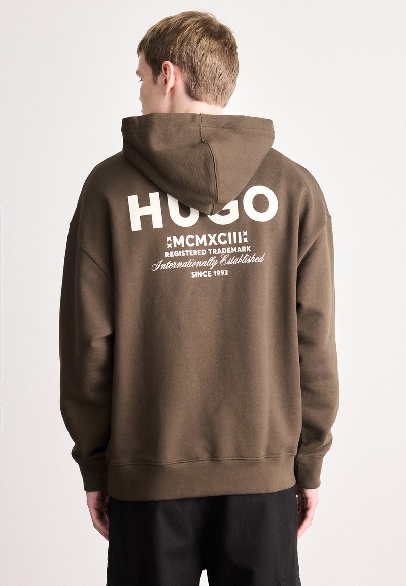 Ruda džemperis su gobtuvu, kurio priekyje yra didelis baltas užrašas „HUGO“ ir prekės ženklo detalės gale. Minkšta tekstūra ir laisvas siluetas.