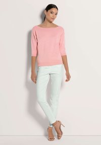 Frau trägt einen pinkfarbenen Pullover mit Dreiviertelärmeln, weiße enganliegende Hose und nudefarbene High-Heelsandalen, während sie vor einer hellen Wand steht.