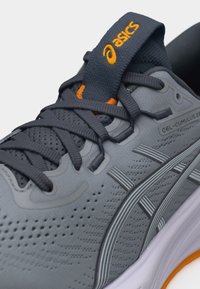 Γκρι παπούτσι τρεξίματος ASICS Gel-Cumulus 26 με σκούρα γκρι κορδόνια, αναπνεύσιμο επάνω μέρος από δίχτυ και πορτοκαλί λογότυπα στη γλώσσα και τη σόλα.