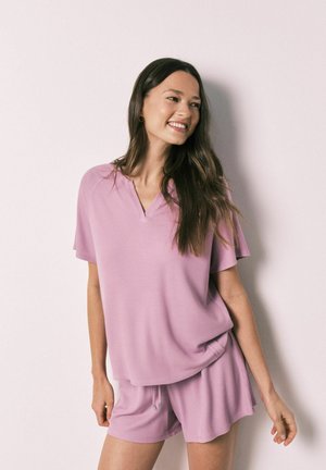 Kvinde med langt brunt hår iført en løs lavendel kortærmet top og matchende shorts, smilende og stående op ad en lyse væg.