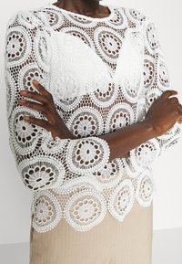 Haut en crochet blanc avec des motifs floraux, un col rond et des manches longues, associé à un bas en tissu beige. Design texturé et transparent.
