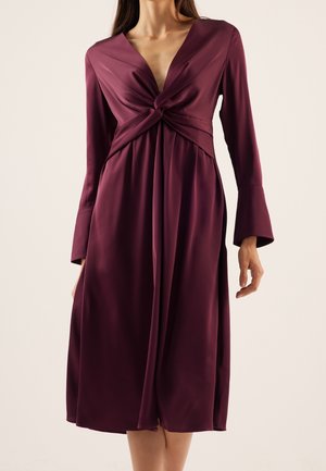 Femme portant une robe midi à manches longues, violet profond, avec un détail noué devant et un décolleté en V sur un fond uni.