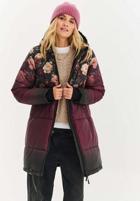 Prošívaná bunda s květinovým vzorem nahoře, vínově červená spodní část, zipové zapínání a kapuce. Kombinována s béžovým svetrem a černými kalhotami.