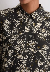 Czarny shirt z białym wzorem kwiatowym i zielonymi akcentami. Posiada kołnierzyk i zapięcie na guziki, wykonany z gładkiego materiału o lekkim połysku.