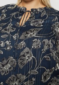 Blusa navy con motivo floreale bianco, scollo elastico con dettaglio a laccio e righe verticali dorate, realizzata in tessuto leggero.
