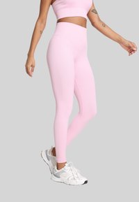 Kobieta ubrana w jasnoróżowe legginsy z wysokim stanem oraz dopasowany top, w białych i szarych butach sportowych, stoi z jedną nogą skrzyżowaną.