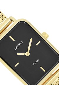 Montre rectangulaire dorée avec cadran noir, marquages minimalistes et bracelet en maille. Les caractéristiques incluent une petite couronne et le texte "Vintage" sur le cadran.