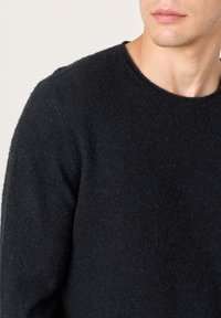 Schwarzer Strickpullover mit rundem Ausschnitt, weicher Textur und langen Ärmeln. Vor hellem Hintergrund präsentiert, konzentriert auf den Oberkörper.