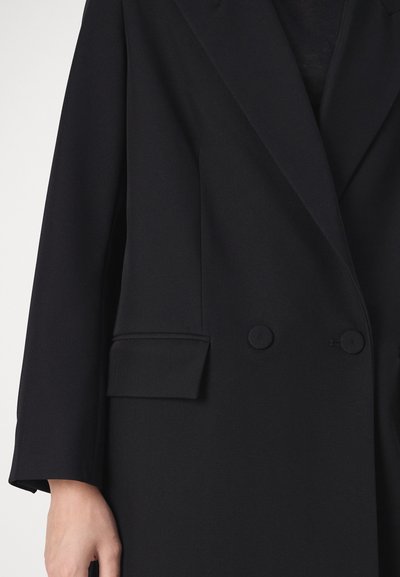 Schwarzer, maßgeschneiderter Blazer mit langen Ärmeln, zwei verdeckten Knöpfen, einer Pattentasche und breiten Revers, getragen über einem schwarzen Top.