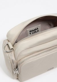 Beżowa torba z teksturowanego materiału, zamykana na suwak, z metką z logo wewnątrz. Charakteryzuje się kompaktowym, prostokątnym kształtem i gładkim wykończeniem.