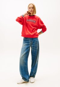 Červená mikina s logom "tommy", voľný strih a žebrované manžety. Kombinovaná s modrými džínsami s širokými nohavicami a bielymi teniskami na svetlom pozadí.