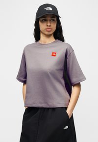 Γυναίκα που φοράει μωβ κοντομάνικη μπλούζα, μαύρη φούστα και μαύρο καπέλο, όλα με το λογότυπο The North Face, στέκεται με τα χέρια στις τσέπες.