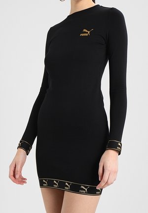 Mujer vistiendo un vestido corto negro ajustado de manga larga con logotipos dorados de Puma en el pecho, puños y dobladillo sobre un fondo liso.