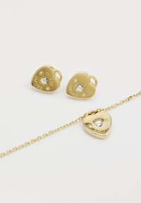 Boucles d'oreilles et pendentif en forme de cœur en or avec une pierre précieuse transparente et le texte gravé "GUESS AMORE", présentant un design de chaîne liée.