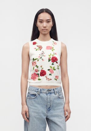 Jeune femme portant un haut blanc sans manches avec broderies florales et papillons roses et rouges et un jean bleu clair taille haute.