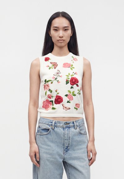 Joven mujer con camiseta blanca sin mangas con bordado de flores y mariposas en rosa y rojo y vaqueros de talle alto en azul claro.