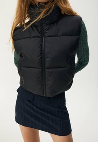 Czarna pikowana kamizelka z wysokim kołnierzem i przednim zamkiem błyskawicznym, w zestawieniu z ciemną, paskowaną spódnicą. Charakteryzuje się gładką, błyszczącą fakturą.