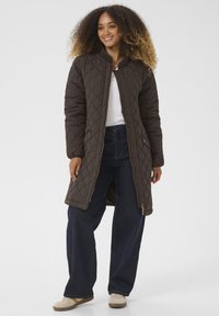 Veste long en cuir marron matelassée avec un col montant, fermeture éclair et poches latérales. Assortie avec un jean taille large foncé et des chaussures beiges.