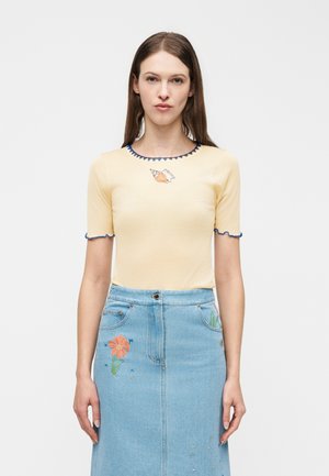 Donna con capelli castani lisci che indossa una maglietta a maniche corte di colore giallo pallido con rifiniture blu e motivo a conchiglia, abbinata a una gonna di jeans azzurro chiaro con ricami floreali.