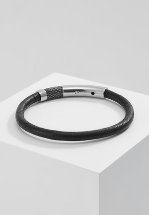 Bracelet en cuir tressé noir avec fermoir magnétique en argent posé sur un socle blanc sur fond gris.