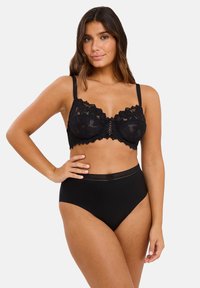 Woman wearing a black lace bra and high-waisted black underwear, standing with one hand on her hip against a white background.